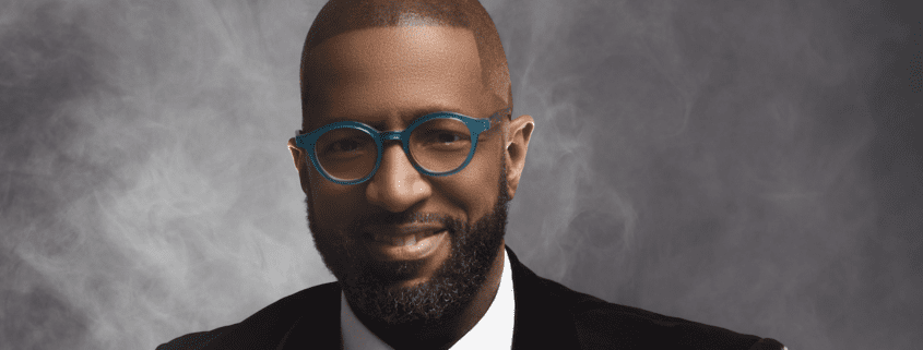 Man wearing glasses smiles against smoky background.